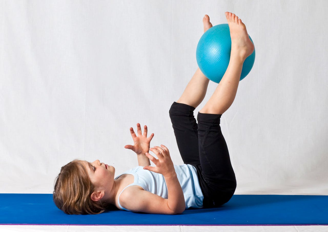 about-us-img Young girl practicing balance exercises with a blue yoga ball indoors on a mat.